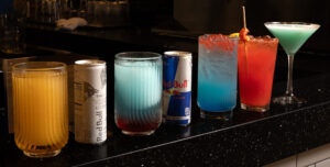 Colorful cocktails and a Red Bull can on a dark bar counter with beer taps in the dim background.