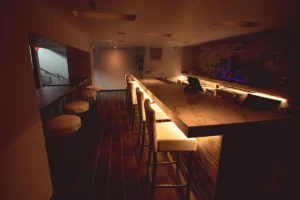 Dimly lit bar interior with a long marble counter and cream bar stools, warm under-counter lighting, and a row of seating along one side.