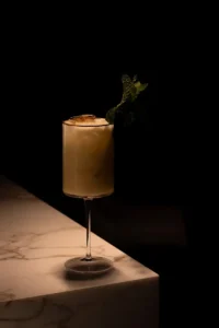 Pale cocktail in a tall stemmed glass with a mint sprig garnish on a marble bar against a dark background.