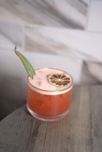 Pink cocktail in a clear glass with a dried lime wheel garnish and a curved green garnish, set on a wooden table with a blurred background.