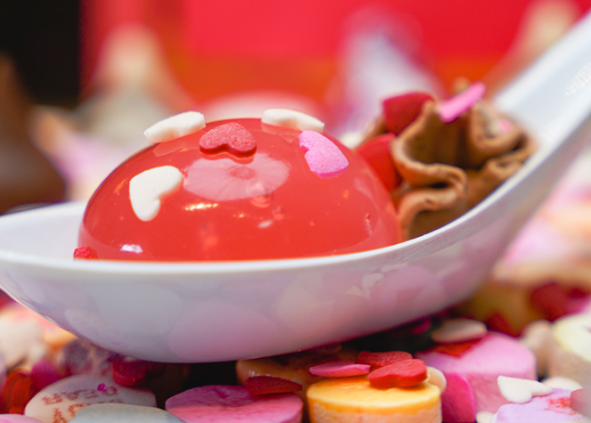 Pink glossy dome dessert with heart-shaped sprinkles on a white spoon, surrounded by colorful candy bits