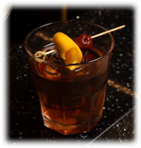 Short tumbler with a dark cocktail on a black speckled bar, garnished with an orange peel twist and a cherry on a skewer