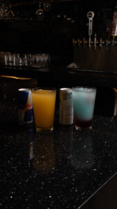 Row of drinks on a dark bar: a Red Bull can, a yellow iced drink, a Coconut Edition Red Bull, and a pale blue cocktail.