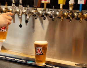 A hand pours beer from a tap at a row of draft beer handles, with an Arizona logo on the glass and a finished glass on the counter nearby.