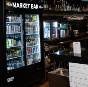 Bar with a glass-door fridge stocked with canned drinks and bottles, a market bar sign above, and rows of liquor bottles on shelves behind the counter.