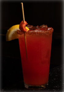 Red cocktail in a sugared rim glass with a lemon wedge and a skewer on a dark bar surface.