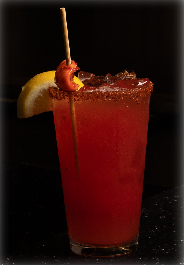 Red cocktail in a sugared rim glass with a lemon wedge and a skewer on a dark bar surface.