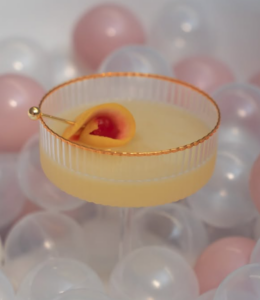 Pale yellow cocktail in a ribbed glass with a gold rim, garnished with a lemon twist and cherry, against a soft pastel balloon backdrop.