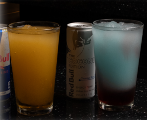 Three beverages on a dark bar counter: a Red Bull can on the left, a tall yellow drink in a glass, a Coconut Edition Red Bull can in the center, and a blue-tinted cocktail with ice on the right.
