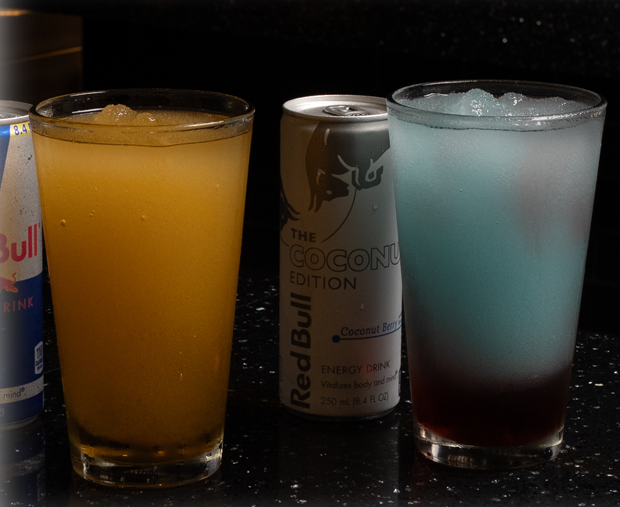 Three beverages on a dark bar counter: a Red Bull can on the left, a tall yellow drink in a glass, a Coconut Edition Red Bull can in the center, and a blue-tinted cocktail with ice on the right.