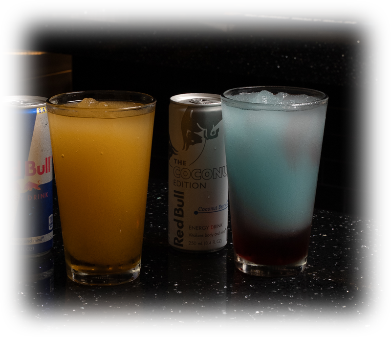 Three beverages on a dark bar counter: a Red Bull can on the left, a tall yellow drink in a glass, a Coconut Edition Red Bull can in the center, and a blue-tinted cocktail with ice on the right.