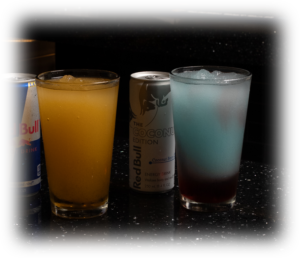 Three beverages on a dark bar counter: a partially visible Red Bull can on the left, a tall glass filled with an orange drink, and a white can labeled 'The GoConu Edition' between two other drinks; on the right is a blue-to-red layered cocktail in a glass with ice.