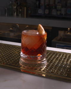 Cocktail in a cut-glass tumbler with a lemon twist garnish on a bar counter, ice-filled and illuminated by bar lights