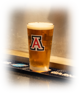 Pint of beer with a foamy head on a bar, the glass featuring a large red and white A logo.
