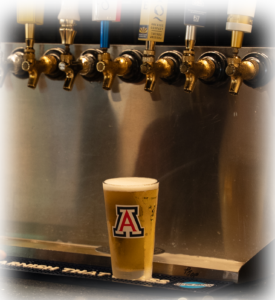 Amber beer in a tall glass with an Arizona logo, in front of a row of beer taps behind it.