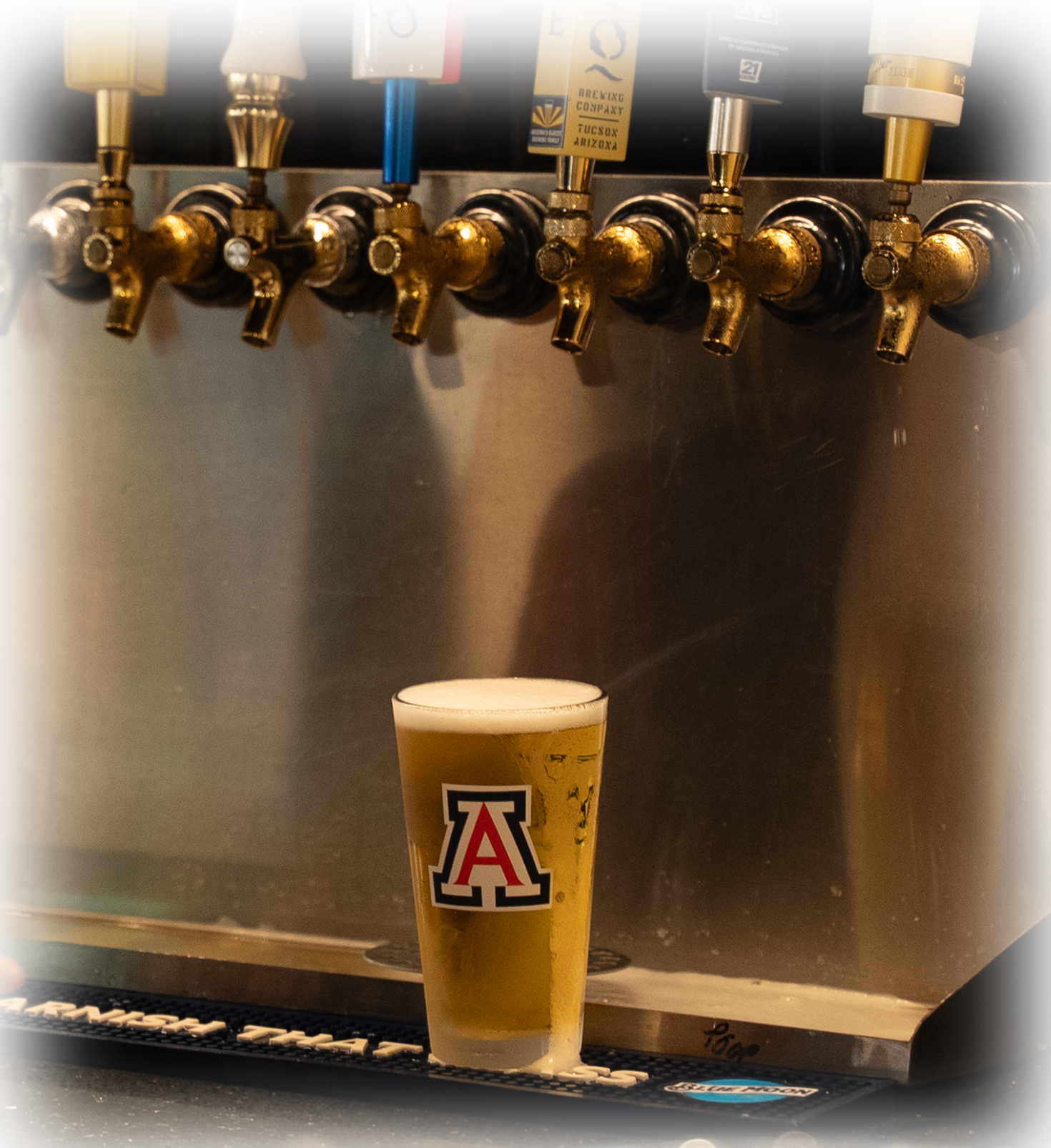 Pint of beer with a foamy head on a bar, the glass featuring a large red and white A logo.