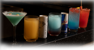 Assorted drinks lined up on a dark bar counter: blue martini, amber tumbler, Red Bull can, gradient blue punch, another Red Bull can, a blue cocktail, and a red fruit punch with a skewer.