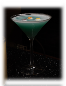 Blue-green cocktail in a martini glass with three small floating garnishes on a dark bar backdrop.