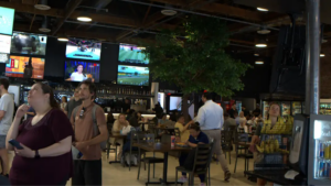 Busy indoor dining area with people seated at tables, a large indoor tree, and multiple TV screens above the bar area.