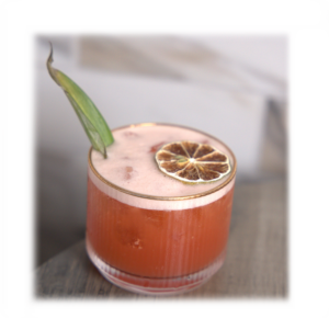 Pink cocktail in a short glass with a lime garnish and a dried citrus wheel, set on a wooden surface.