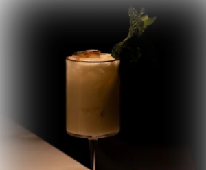 Cocktail with a mint garnish in a stemmed glass on a marble bar, dimly lit against a black background.