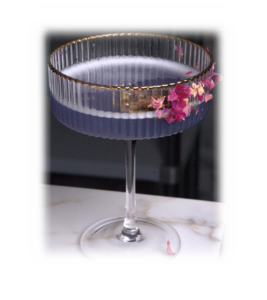 Cocktail in a ribbed coupe glass with a purple drink and pink flower garnish on the rim, placed on a marble counter.