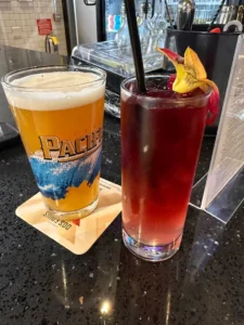 Two drinks on a polished bar counter: a lager in a glass with Pacers branding on a coaster, and a tall red cocktail garnished with fruit and a straw.